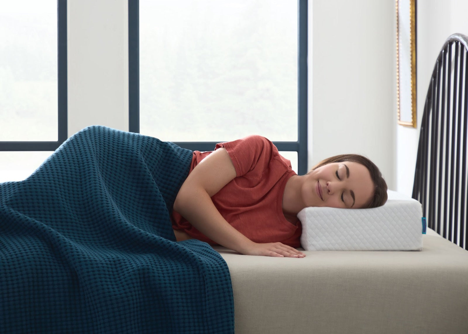 A woman asleep on her side, smiling, her head resting on a Pillow Cube in a bright bedroom.